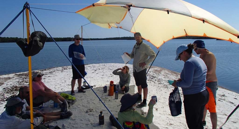 A group of people stand under a tarp shelter on a beach